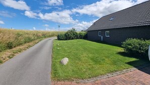 A rural road leading to a dark-colored house with a sloped roof, surrounded by a well-maintained lawn and a field in the background.