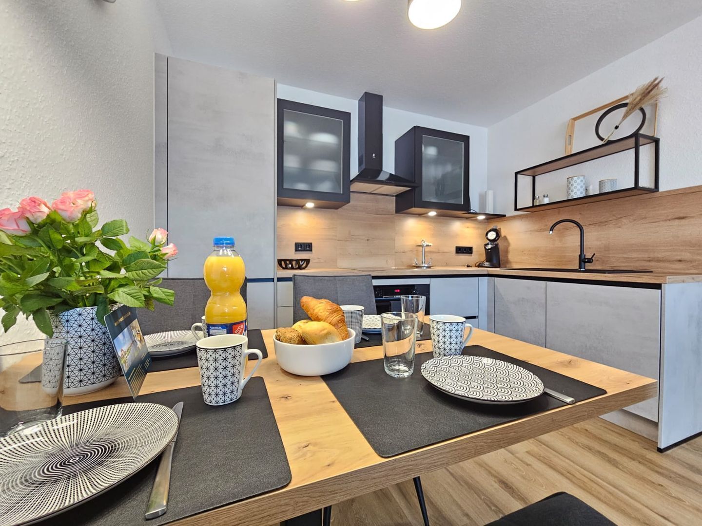 A modern kitchen with a dining area, featuring a breakfast setup with croissants, a bottle of orange juice, and a vase of flowers.