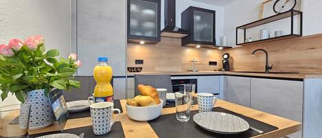 A modern kitchen with a dining area, featuring a breakfast setup with croissants, a bottle of orange juice, and a vase of flowers.