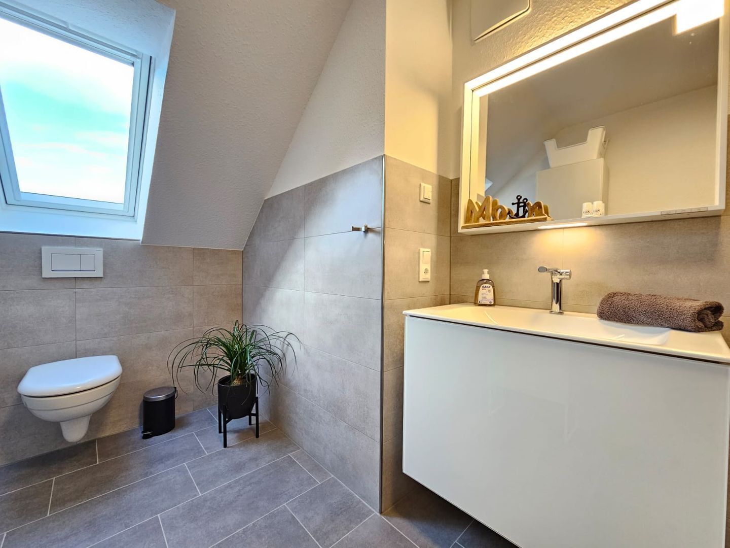 A modern bathroom with a toilet, sink, and mirror. There is a window above the toilet and a plant next to it.