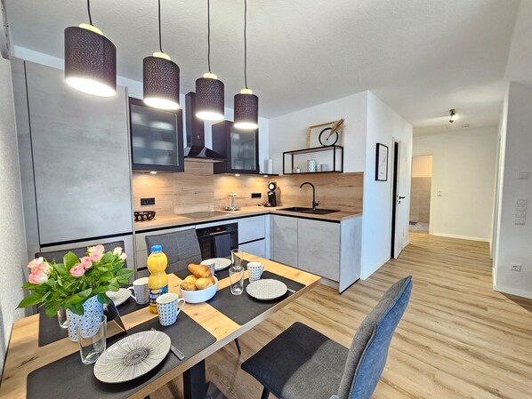 A modern kitchen with pendant lighting, a dining area with a table set for two, and a view into the living room.
