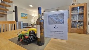 A wooden dining table with a bottle of wine, a fruit basket, and a framed welcome sign.