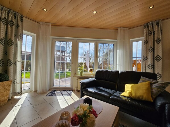 A living room with a black leather sofa, a coffee table with flowers, and large windows with curtains.