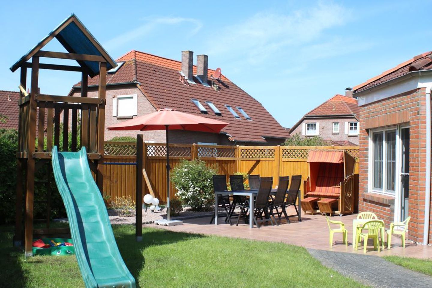 Un patio trasero con un parque infantil, un tobogán, un área de patio con sillas y una casa de ladrillo al fondo.