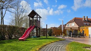 Un parque infantil con un tobogán rojo, una estructura de juego de madera y un camino pavimentado que conduce a una casa con patio.