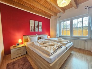 A bedroom with a large bed, wooden headboard, bedside tables, a nightstand, a window with a view, and a red wall with artwork.