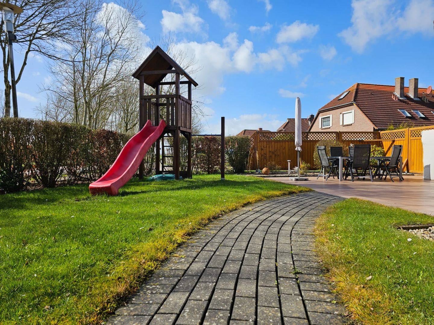 Ein Spielplatz mit einer roten Rutsche, einer Holzspielanlage und einem befestigten Weg, der zu einem Haus mit Terrasse führt.