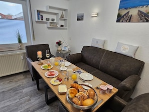 Uma sala de jantar com mesa de madeira posta para o café da manhã, um sofá marrom e vista para a praia pela janela.
