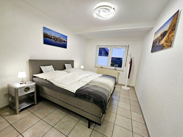 Quarto com cama, mesa de cabeceira, janela e duas imagens penduradas na parede.