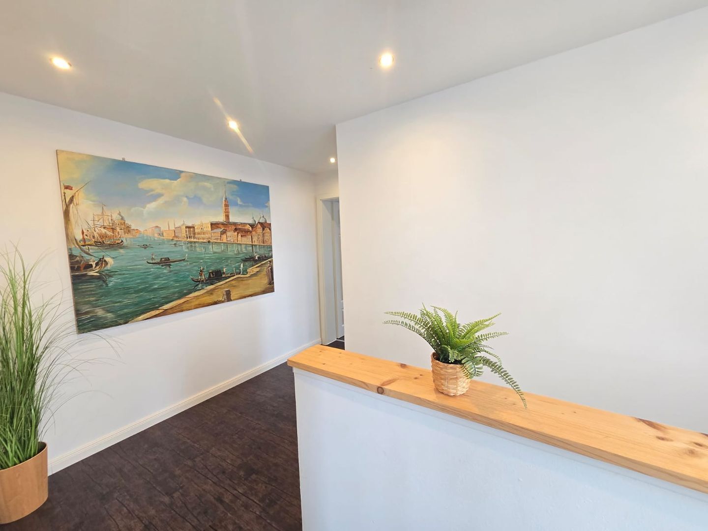 Een receptie met een schilderij van een havengezicht, een houten receptiebalie en een kamerplant.