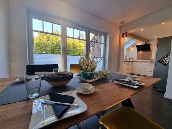 A dining area with a wooden table set for two, a potted plant, and a kitchen in the background.