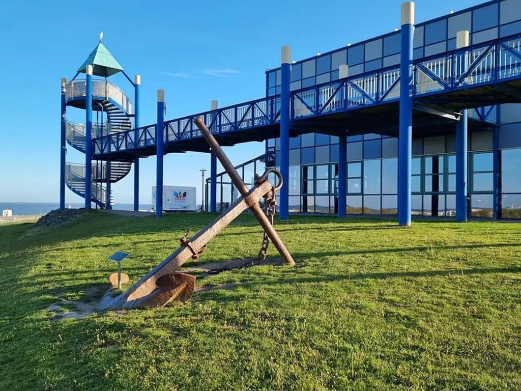 Un parque infantil con un tobogán en espiral y columpios, un edificio azul con fachada de vidrio y un cielo azul despejado.