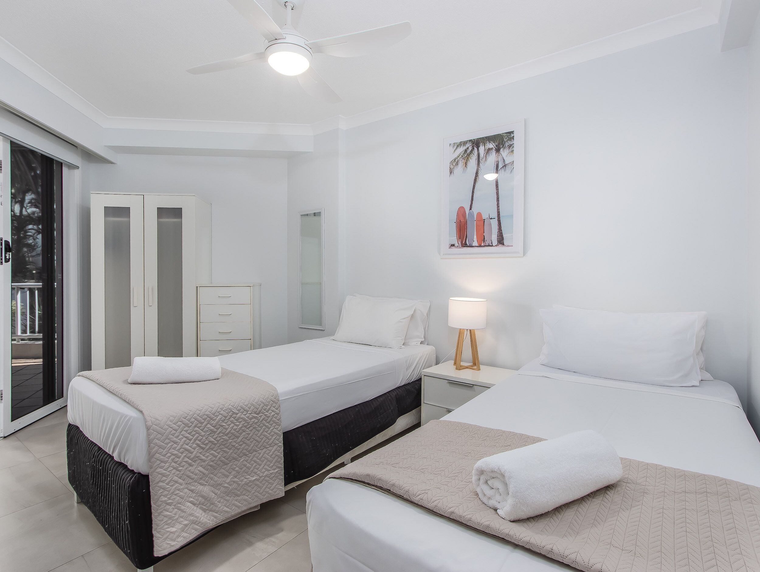 Une chambre d’hôtel avec deux lits, un ventilateur de plafond, une table de chevet avec une lampe, une armoire et une porte-fenêtre coulissante donnant sur le balcon.