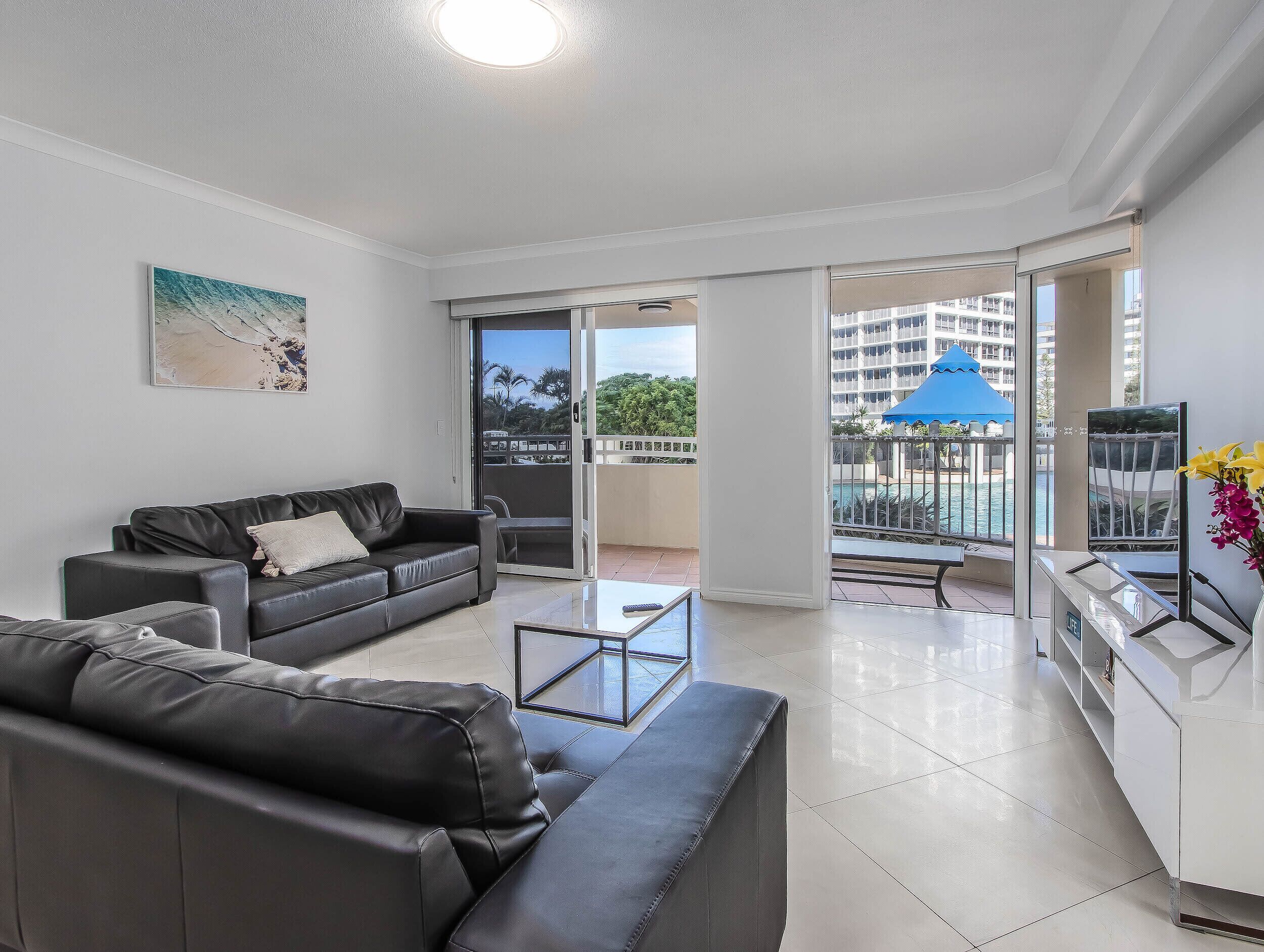 A modern living room with a black leather sofa, a glass coffee table, and a view of a pool and buildings through large sliding glass doors.