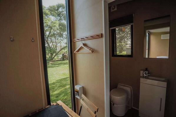 A bathroom with a toilet, a sink, and a mirror. There is a wooden hanger on the wall and a window providing a view of the outdoors.