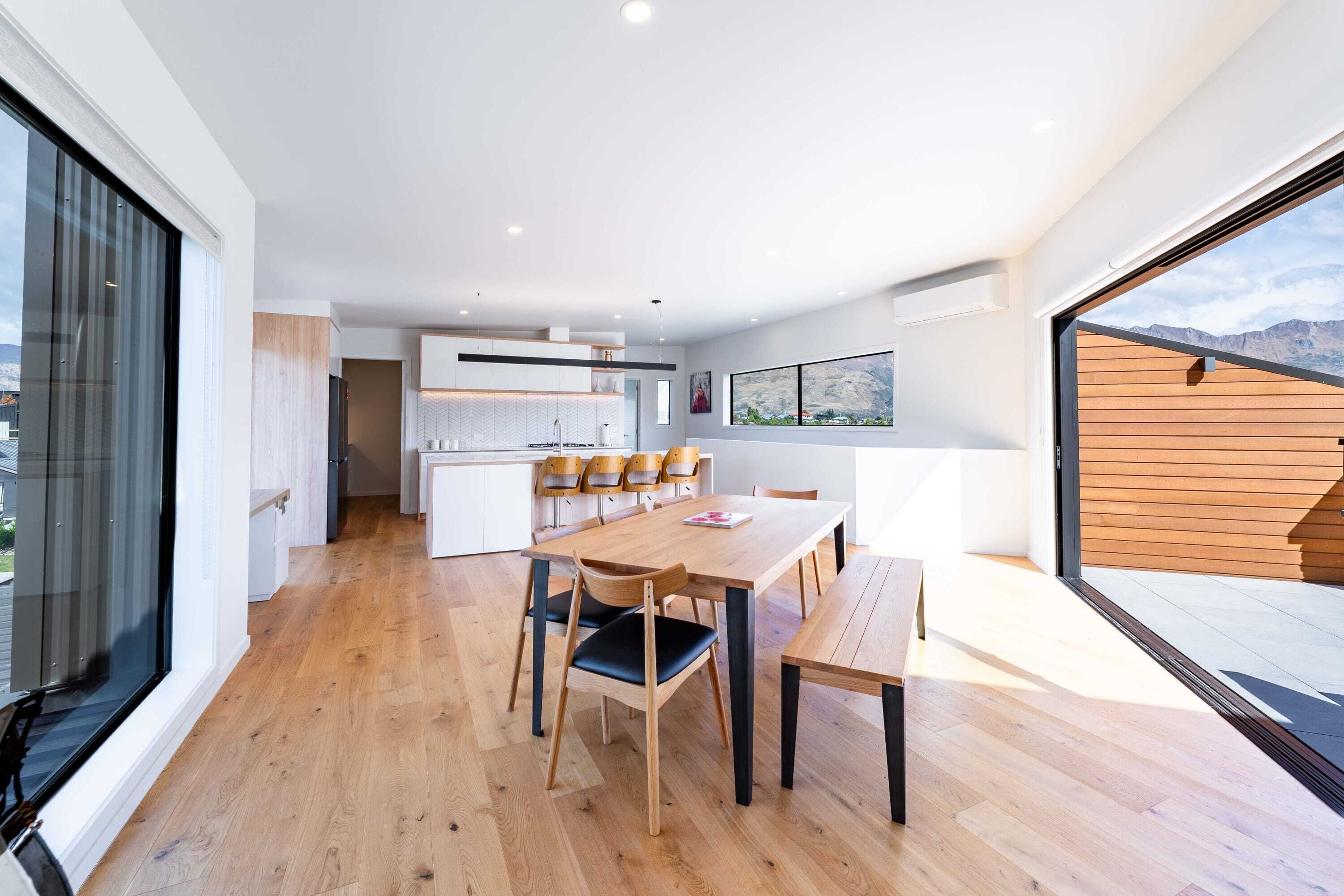 A modern kitchen with wooden cabinets and a large island, a dining area with a wooden table and chairs, and a large window offering a view of the mountains.