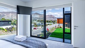 A modern bedroom with a large bed, a view of a cityscape, and a sliding glass door leading to a balcony. - Grand Alpine Vista Escape (Wanaka)