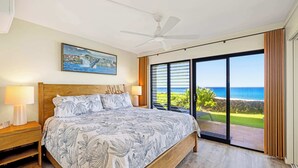 Un dormitorio con cama, ventilador de techo, mesita de noche, lámpara, un cuadro y vista al océano.