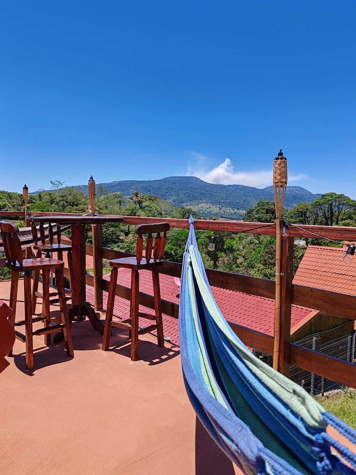 A wooden deck with a hammock, chairs, and a table overlooking a mountainous landscape.