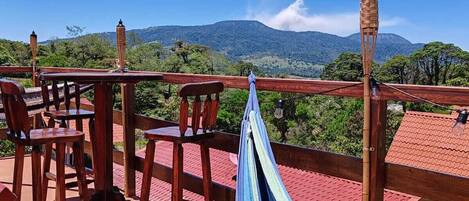 A wooden deck with a hammock, chairs, and a table overlooking a mountainous landscape.