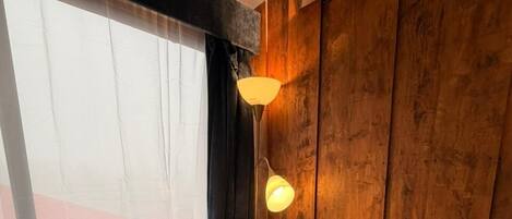 Un coin repas avec une table ronde en bois, deux chaises blanches, un luminaire suspendu moderne et une vue par une fenêtre ornée de rideaux légers.