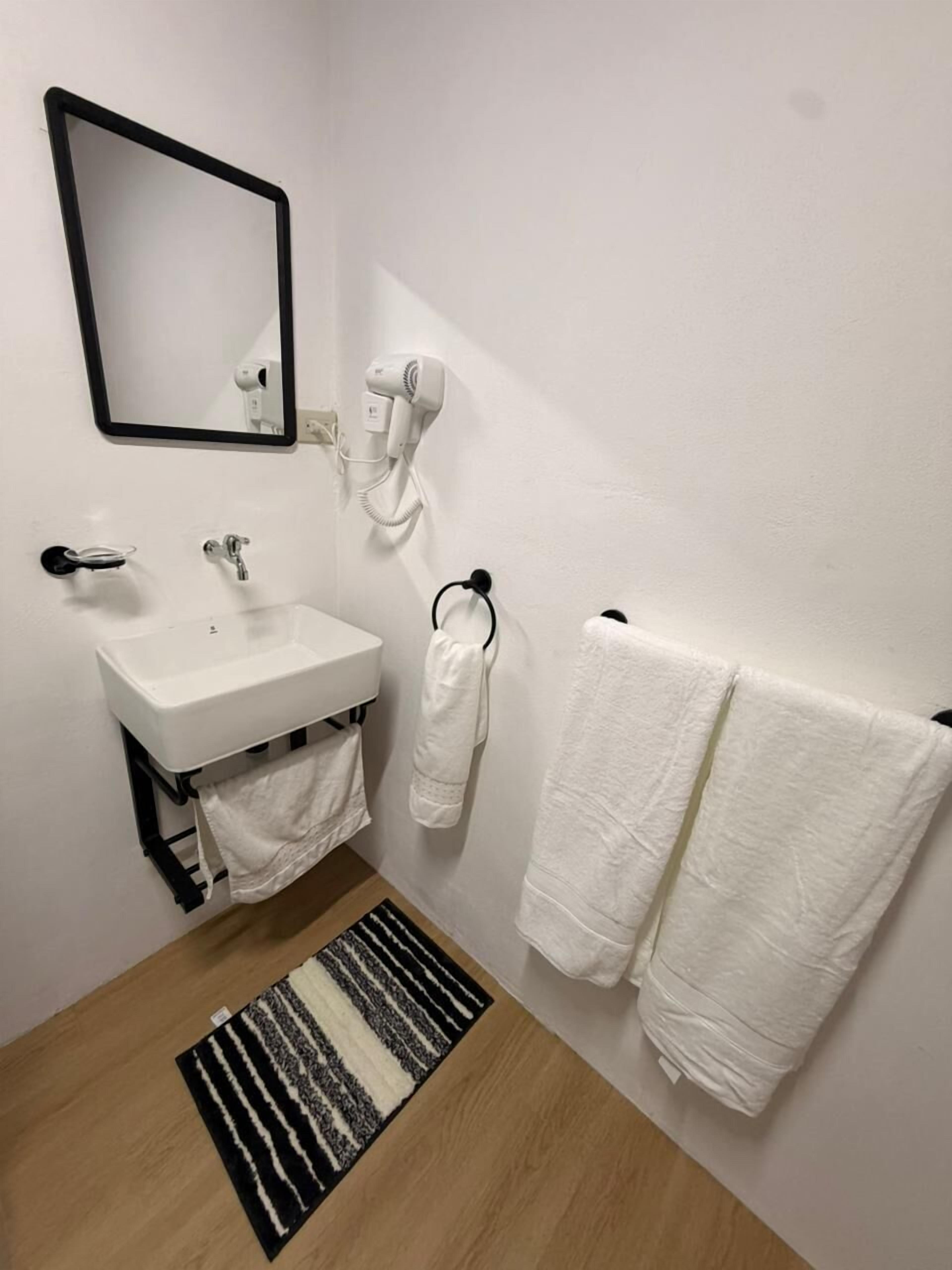 Un baño con un lavamanos blanco, un secador de pelo, un perchero con dos toallas, un espejo y un felpudo a rayas.