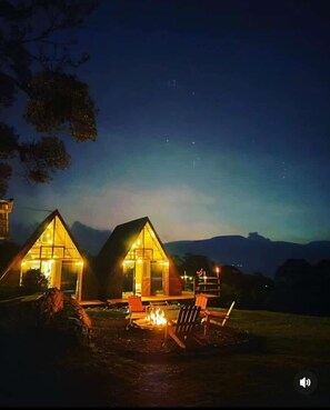 Dos cabañas iluminadas con un pozo de fuego al frente, frente a un paisaje montañoso.