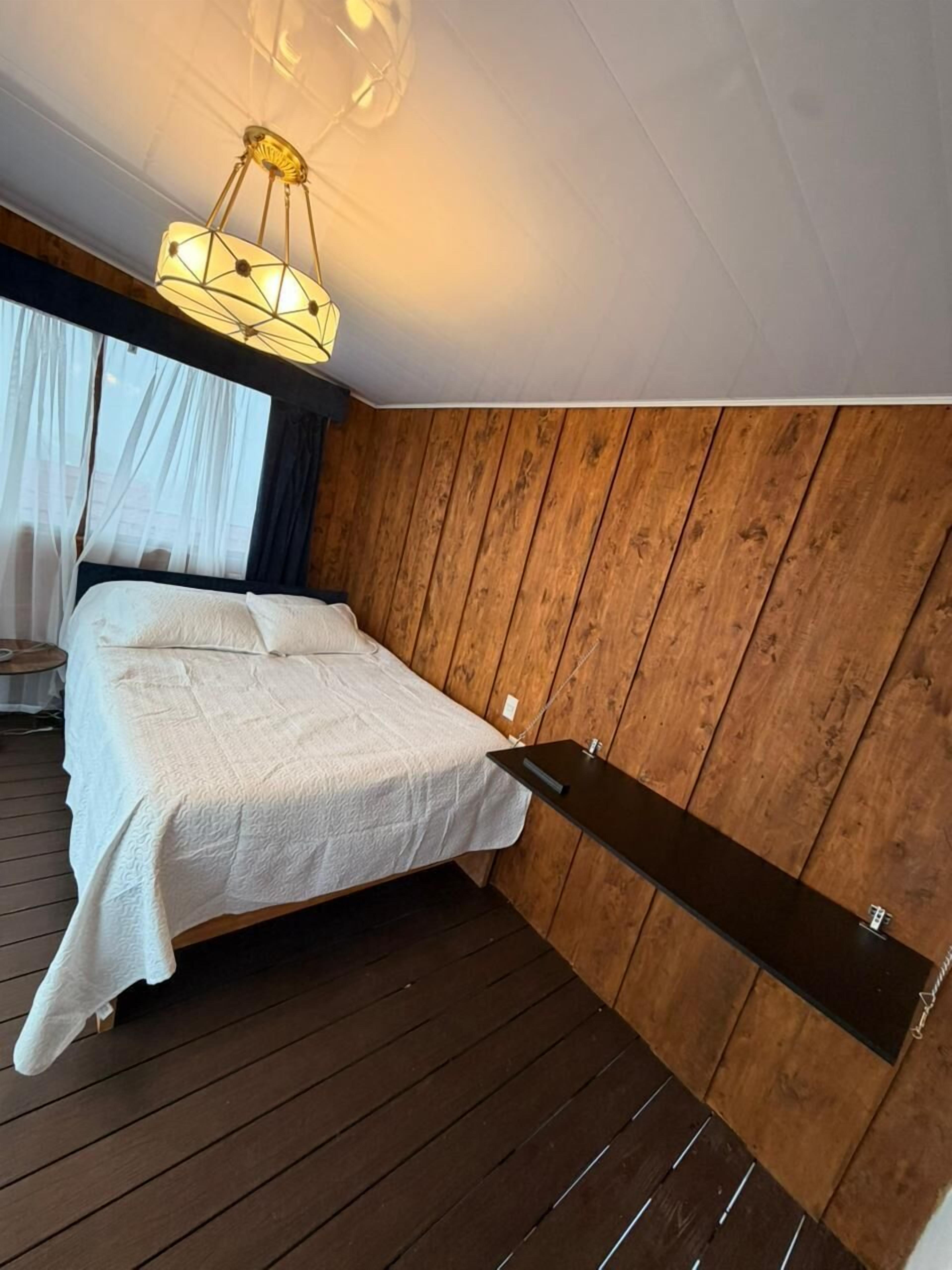 A bedroom with a bed, a hanging light fixture, wooden walls, and a window with curtains.