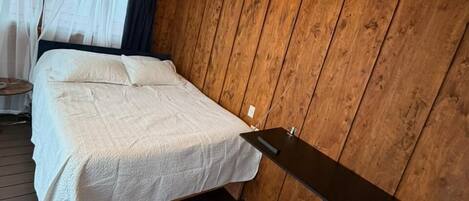 A bedroom with a bed, a hanging light fixture, wooden walls, and a window with curtains.