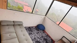 A room with a sofa, a blue cushion, a small wooden table, and large windows offering a view of buildings and trees. - Chalets Vistas del Poas (Vara Blanca)