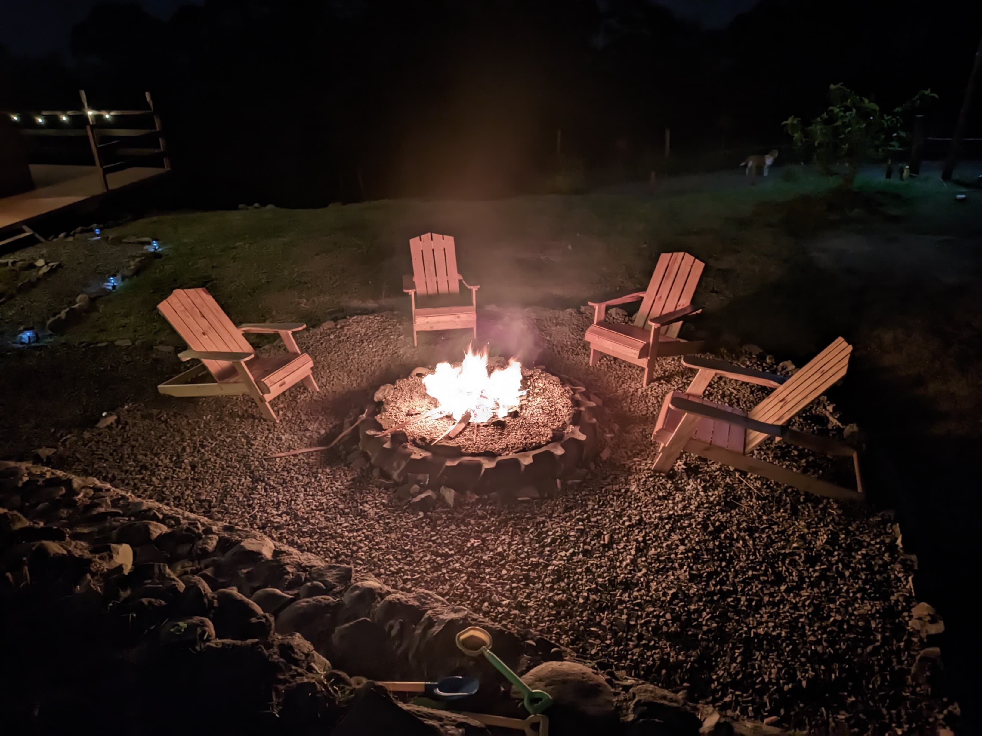 Un área de estar al aire libre iluminada por la noche, con una fogata rodeada de sillas Adirondack de madera.