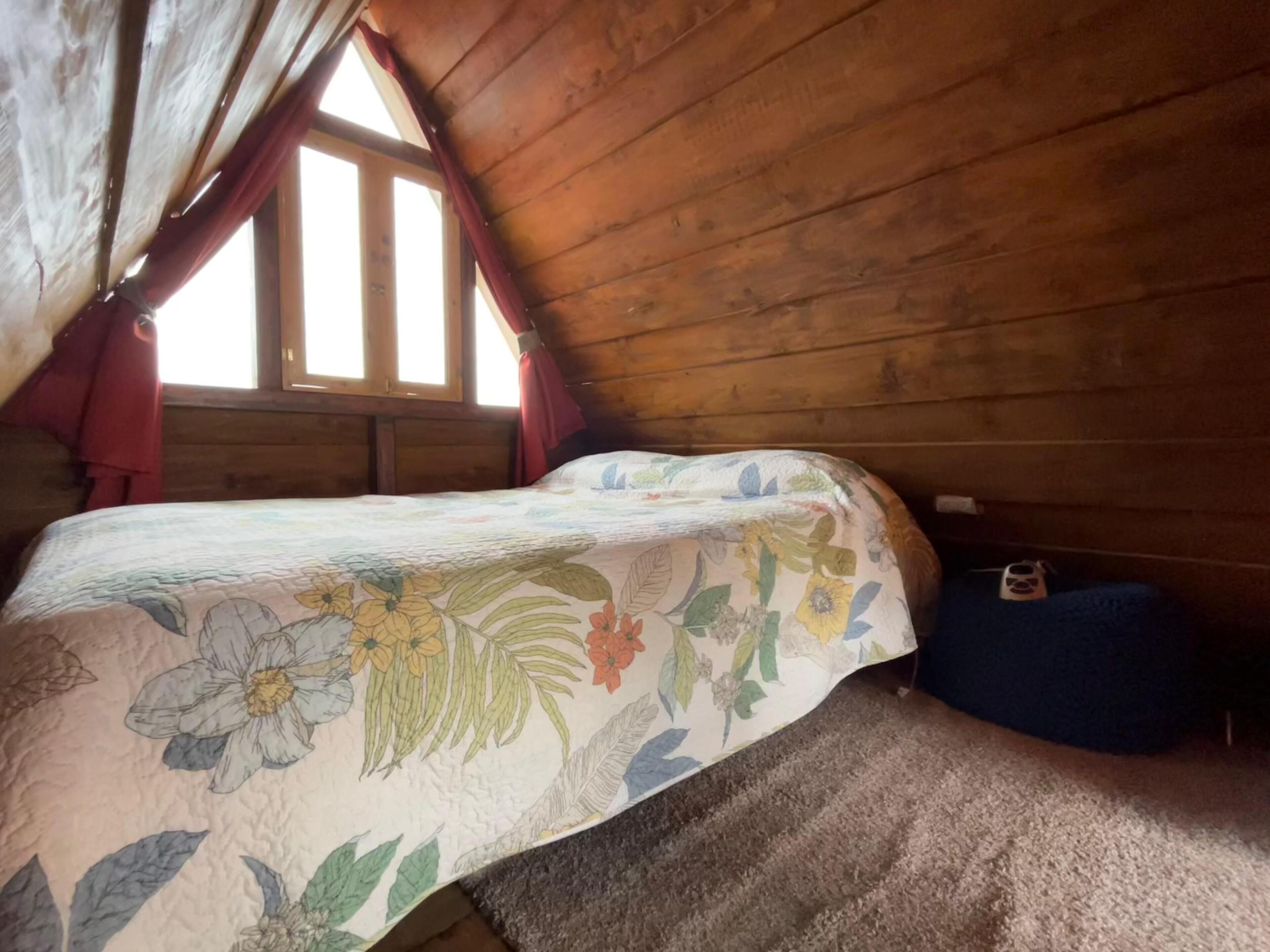 Um quarto de sótão acolhedor com teto inclinado de madeira, uma cama simples com colcha florida e uma pequena cama azul para animais de estimação.