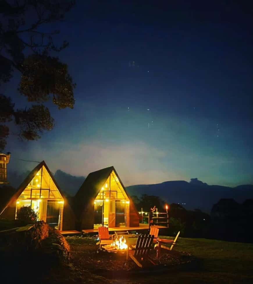 Two illuminated cabins with a fire pit in the foreground, set against a mountainous backdrop.
