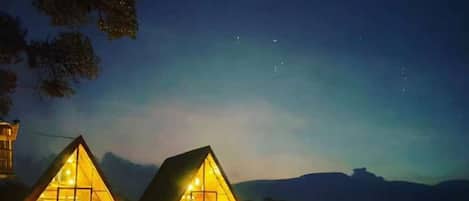 Two illuminated cabins with a fire pit in the foreground, set against a mountainous backdrop.