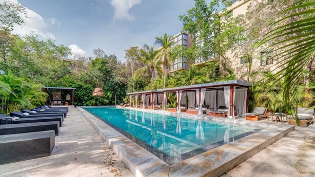 A swimming pool with lounge chairs and a covered seating area, surrounded by lush greenery and a building in the background. - Arthouse Stay with Pool, Gym, & Private Terrace (Tulum)