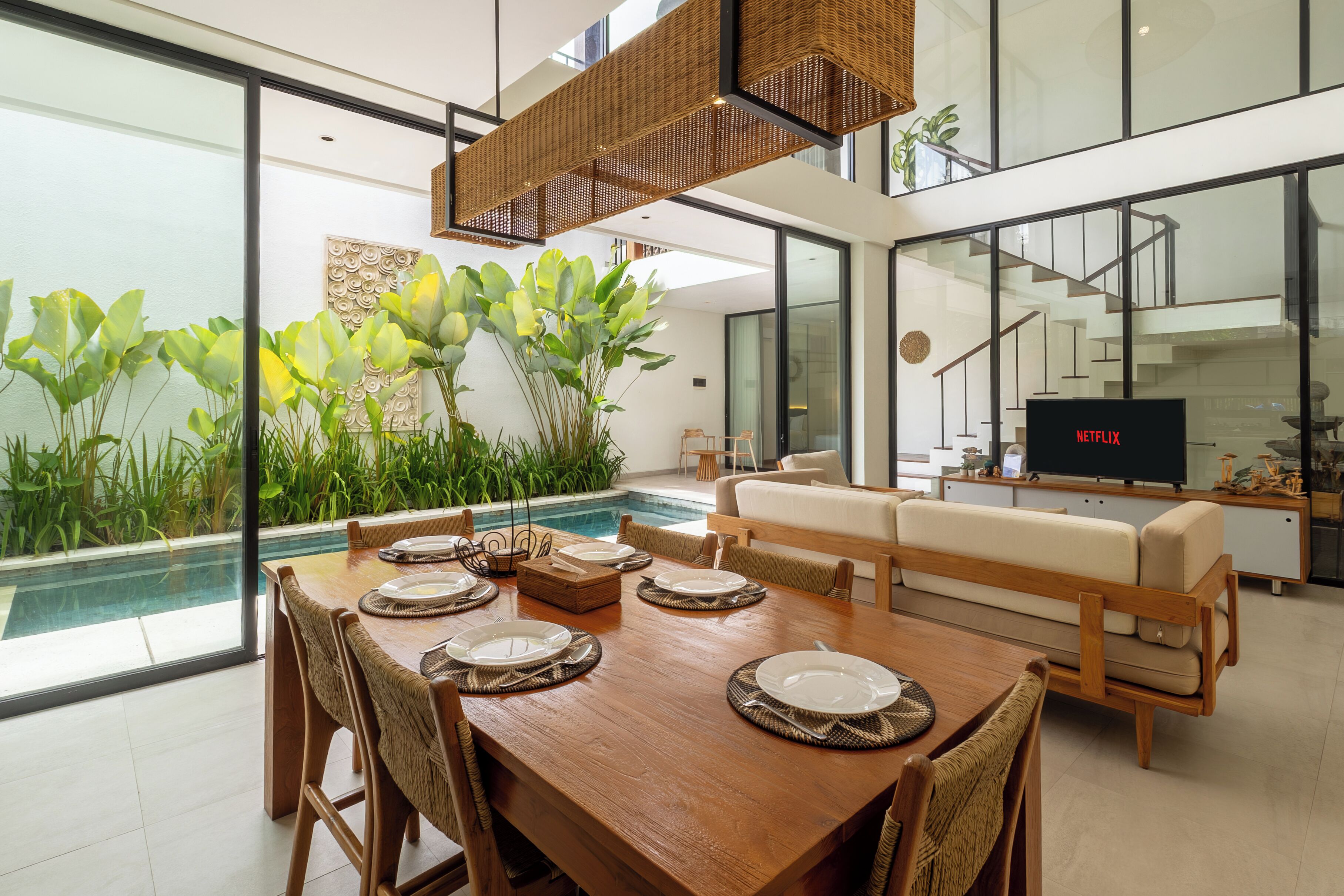 Un comedor moderno con una mesa de madera dispuesta para cuatro personas, un sofá y un amplio ventanal con vistas a una piscina.