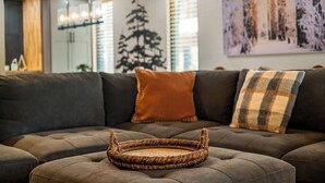 Una sala de estar acogedora con un sofá modular, una bandeja tejida y un cuadro con temática de bosque en la pared.