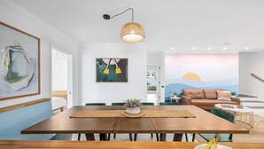 A dining area with a wooden table, chairs, a hanging light fixture, and a wall mural. - Modern 4BR w/ Heated Pool | It's Always Sunny (North Myrtle Beach)