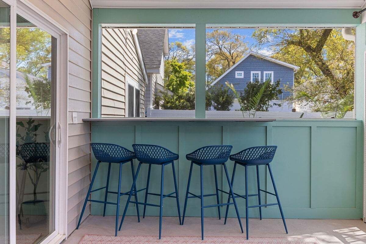 Un porche cubierto con barandilla verde, una ventana con vista a una casa y cuatro taburetes azules.