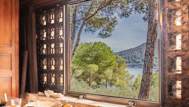 A room with a large window overlooking a lake and trees, a wooden table with glasses and a bowl of fruit, and a wooden door with a decorative metal grille. - Residenza Capo Perla - Capo Perla 2 (Capoliveri)