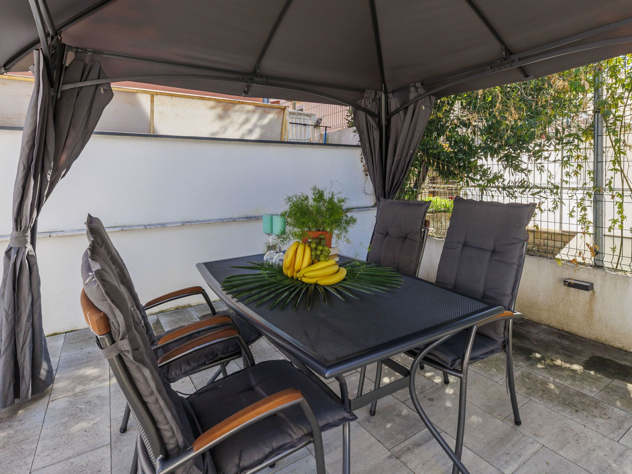 A covered patio area with a table and four chairs, a potted plant, and a bunch of bananas on the table.
