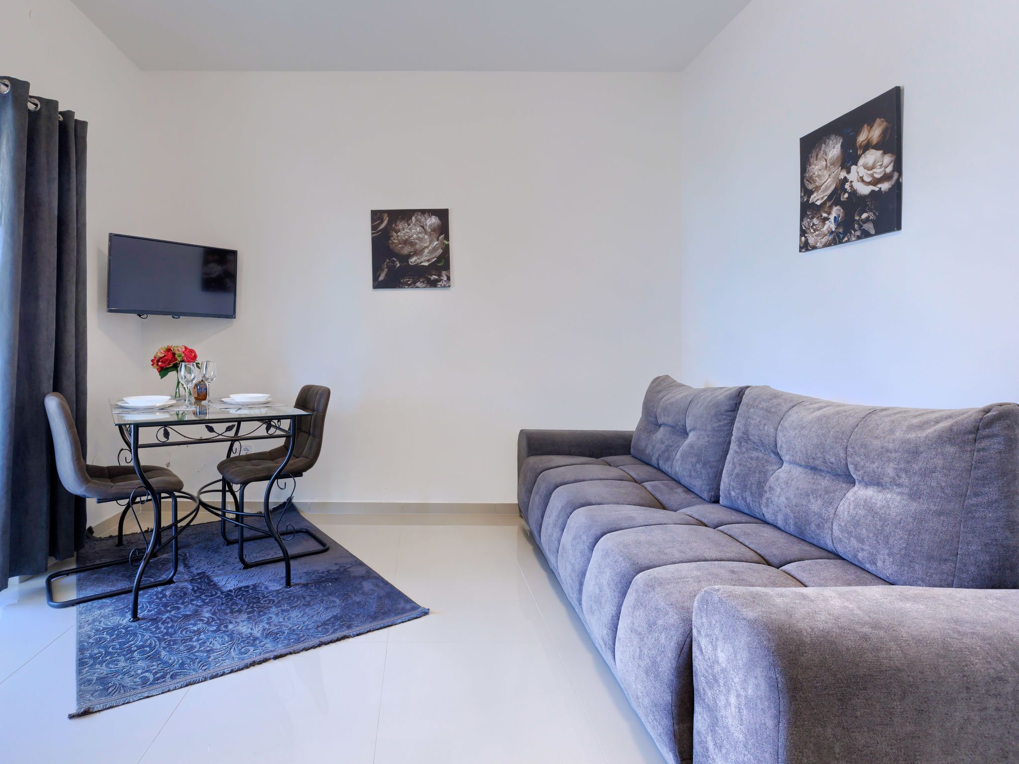Uma sala de estar moderna com um sofá cinzento, uma mesa de jantar com cadeiras, uma TV de tela plana na parede e duas obras de arte emolduradas.