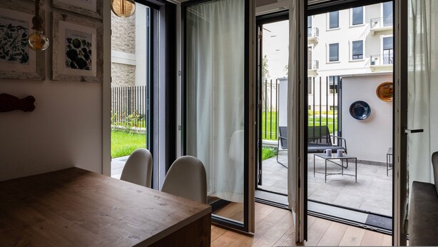 Une chambre moderne avec une table en bois, deux chaises, une porte-fenêtre coulissante donnant sur un patio extérieur équipé d’une chaise et d’une table, et une vue sur un immeuble résidentiel.