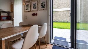 Un coin repas avec une table en bois et des chaises blanches, un micro-ondes rouge, des reproductions botaniques encadrées accrochées au mur et, par une porte vitrée, une vue sur un espace extérieur verdoyant.