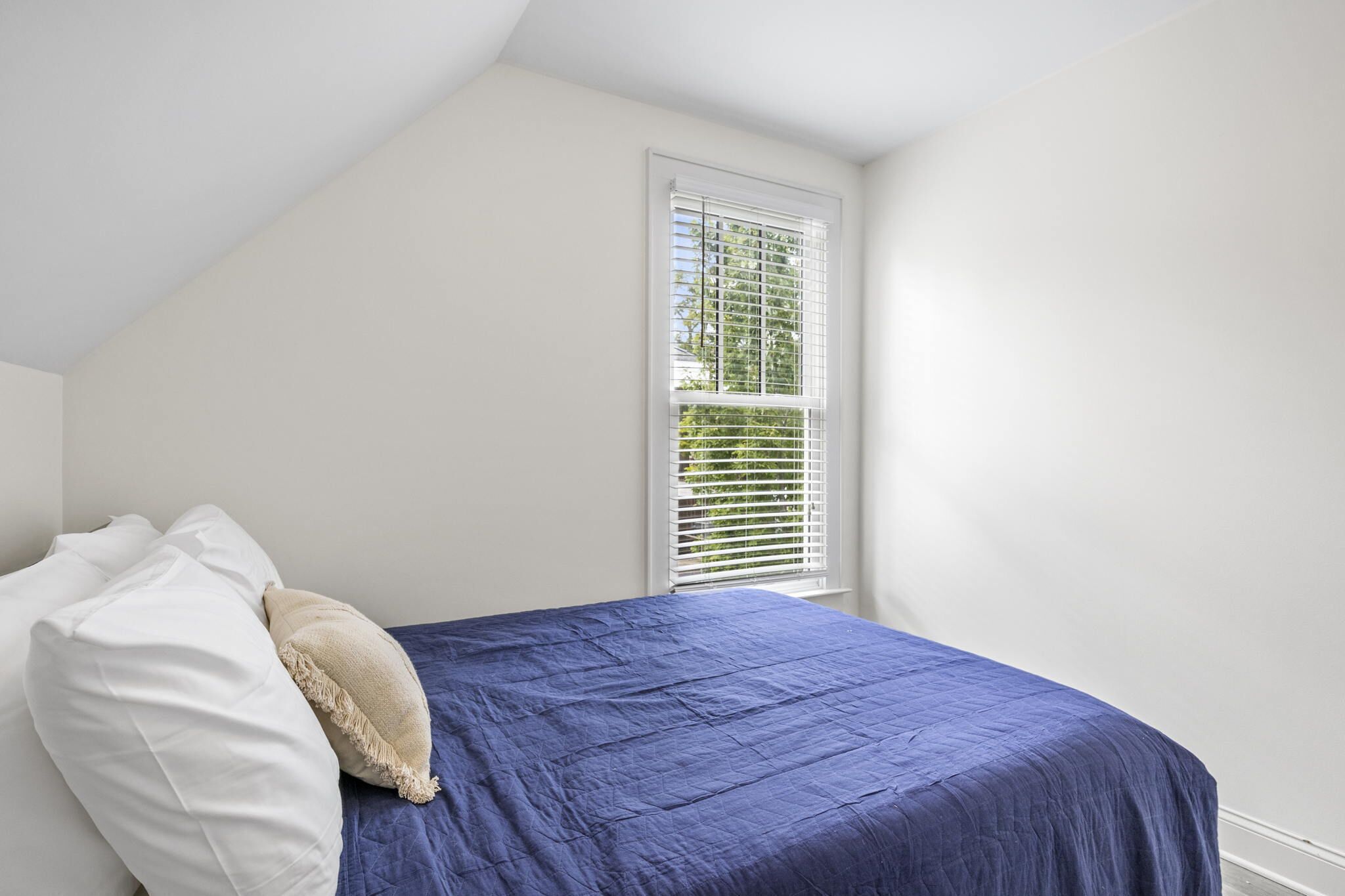 A bedroom with a bed, white walls, a window with blinds, and a view of greenery outside.