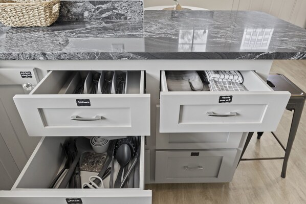 A kitchen with a marble countertop and white cabinetry. There are labeled drawers for utensils, pots, pans, and oven mitts.