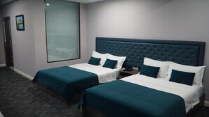 A hotel room with two beds, each with a dark blue bedspread and white linens. There is a large window with a view of a building, and a door to the left. - Bayantsambagarav Hotel (Olgii)