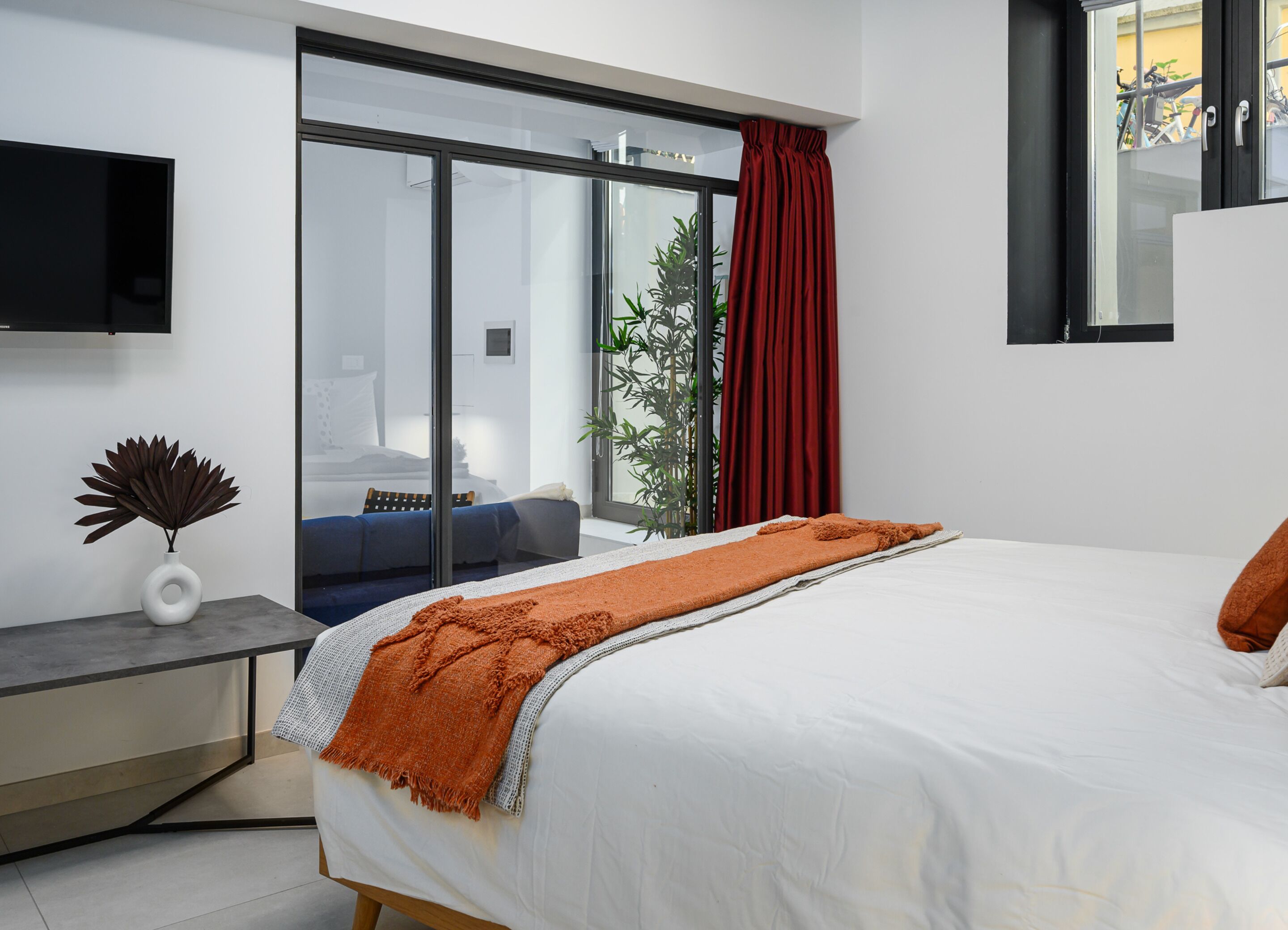 A modern bedroom with a large bed, a TV mounted on the wall, and a sliding glass door leading to a balcony.