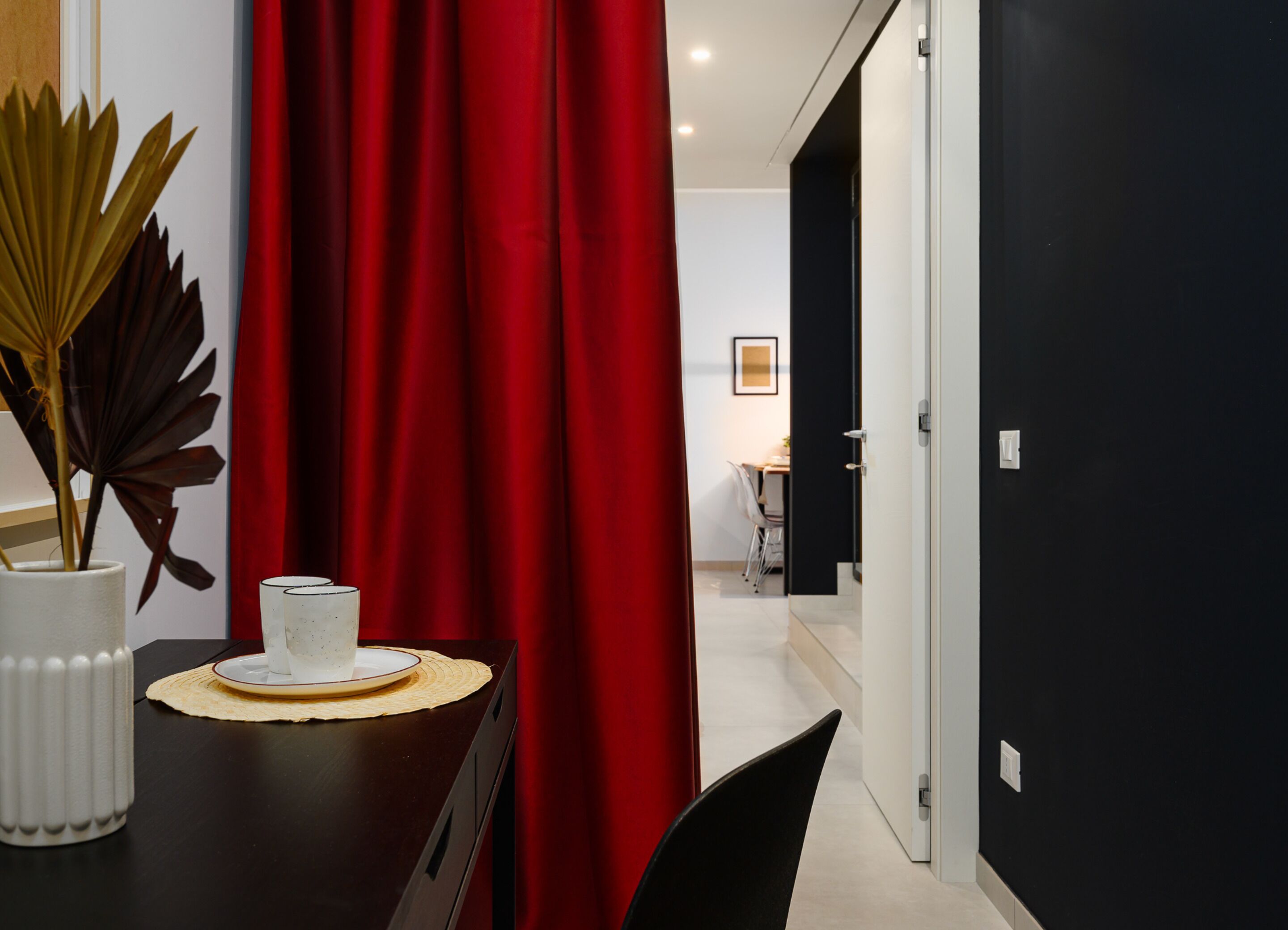 A dining area with a table set for two, a red curtain, and a view into a room with a bed and a chair.