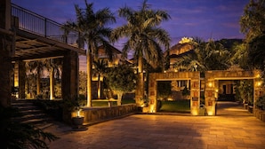 A well-lit courtyard with palm trees, stone architecture, and a view of a hilltop building. - Devi Palace Monsoon Palace View Resort Udaipur (Udaipur)